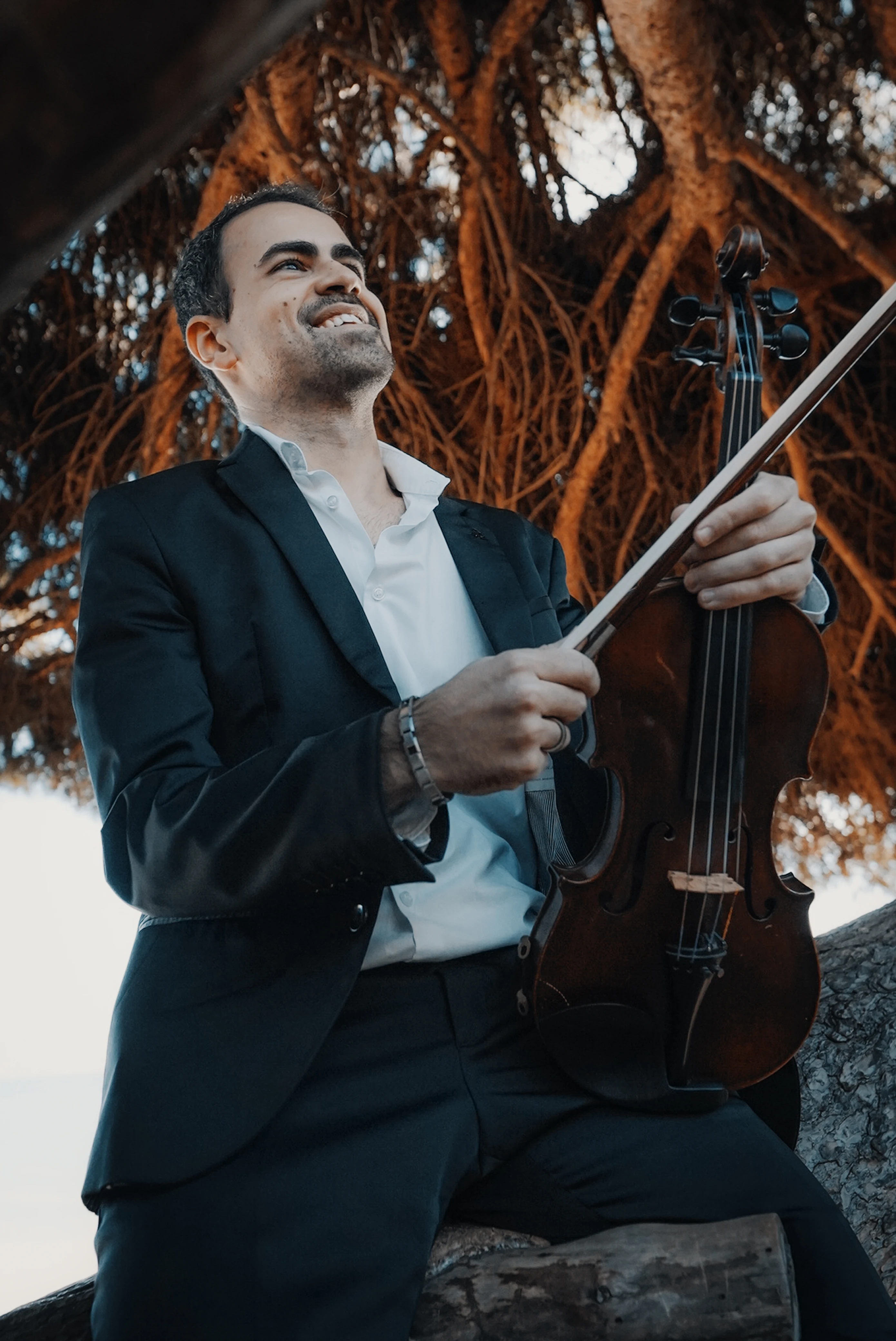 E. Sousa playing violin while seated on a tree beside the sea, wearing a black suit